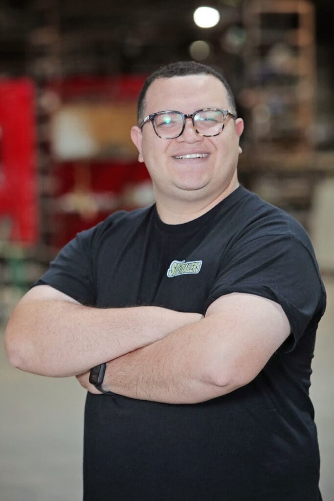 A man with glasses and a black t-shirt smiles with his arms crossed in front of a blurred background.